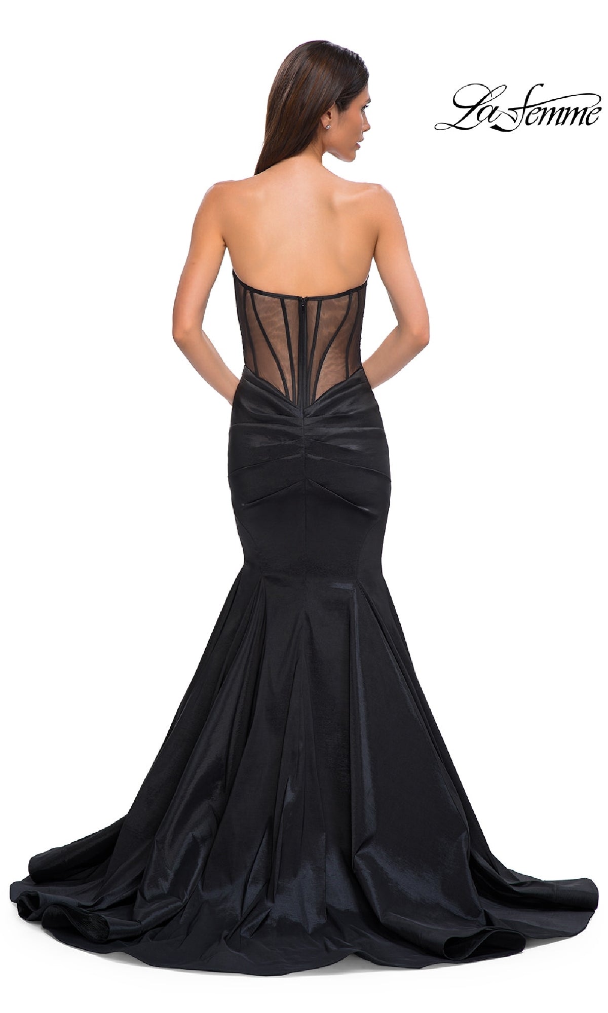 A woman in the Strapless Long Mermaid Prom Dress: La Femme 32791, featuring a fitted black silhouette, sheer back panel, and gathered detailing, stands with her back to the camera, radiating timeless elegance.
